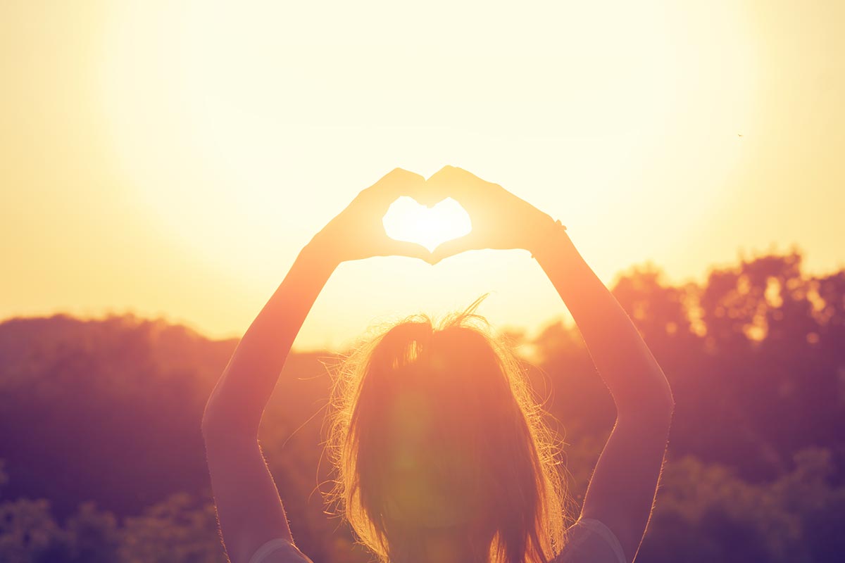 Junge Frau formt Herz mit Händen in den Sonnenuntergang