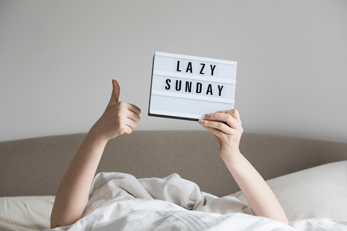 Person im Bett hält Schild mit lustigem Spruch zum Sonntag