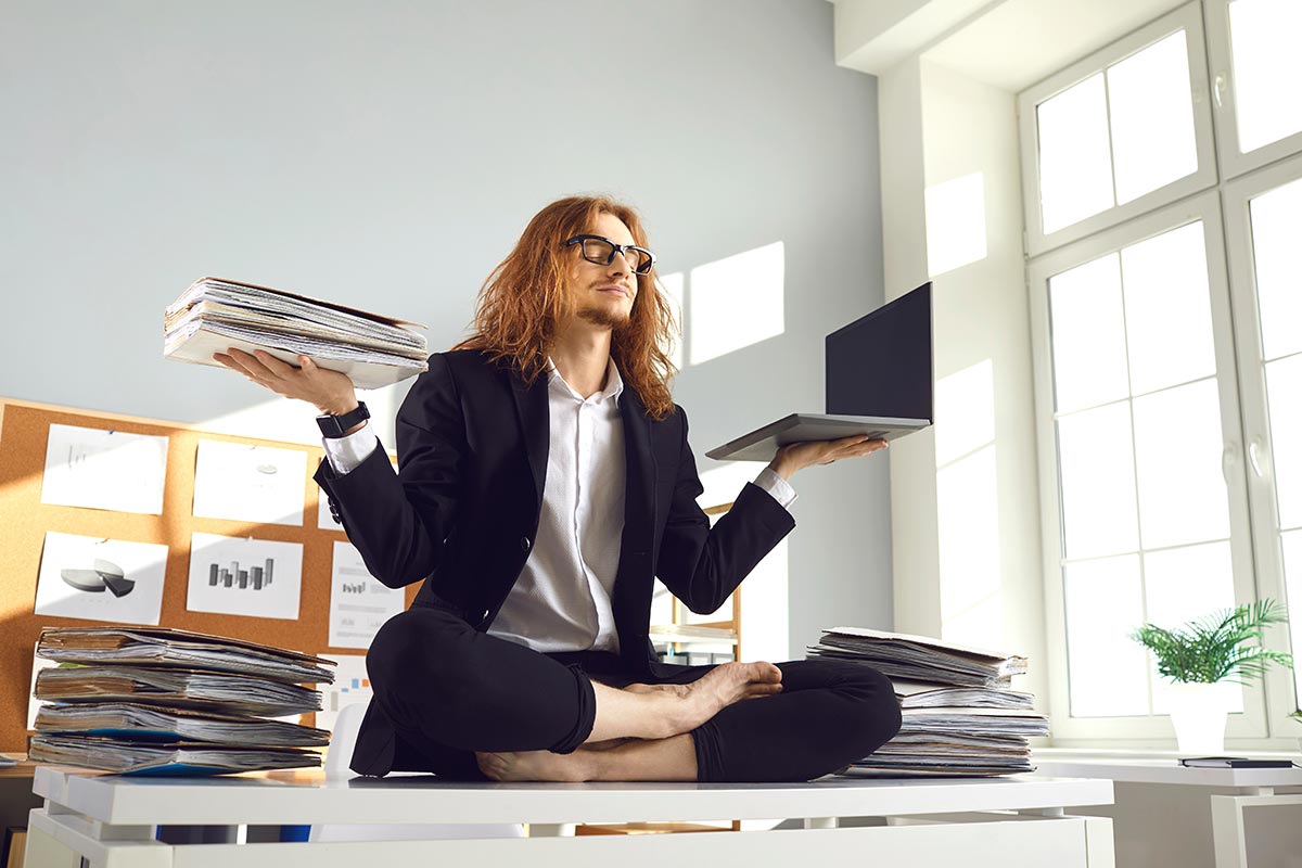 Angestellter in Büro balanciert Laptop und Akten in Yoga Pose