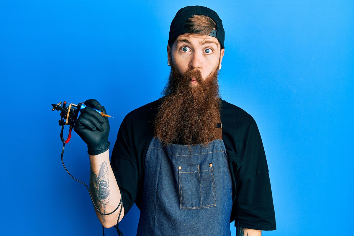 Junger Tätowierer mit langem Bart vor blauem Hintergrund